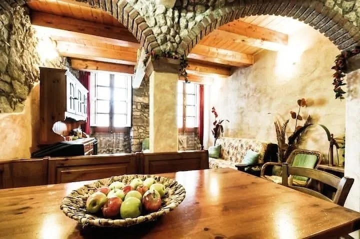 Soggiorno di Su'e Lentu con archi in pietra, travi in legno, divano a fiori e cesto di mele sul tavolo