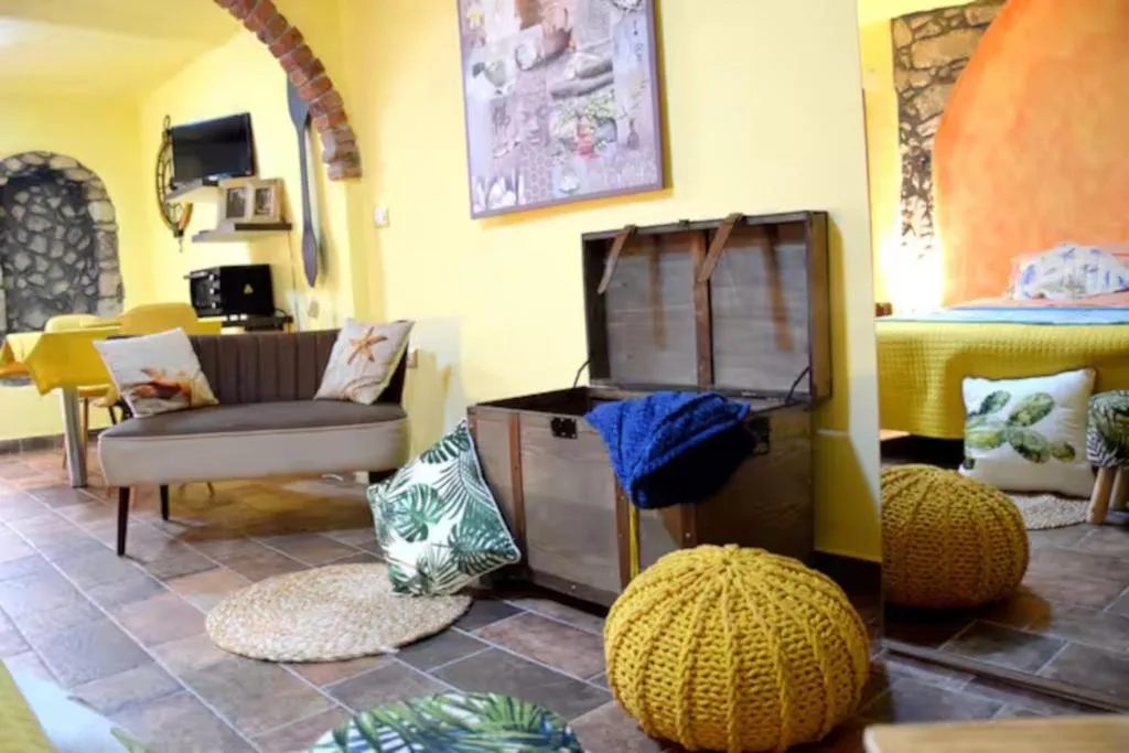 Soggiorno del bilocale Su'e Lentu con pareti gialle, archi in pietra, divanetto, baule in legno e pouf gialli all'uncinetto