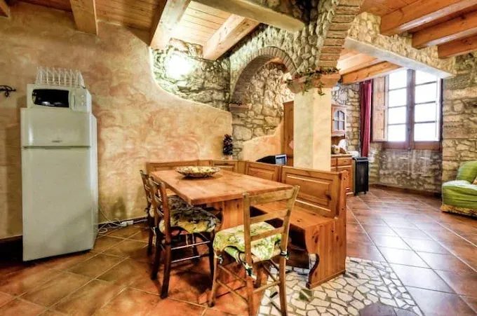 Su'e Lentu, cucina con tavolo e panca lignei, arco tra muri in pietra, soffitto a travi, frigo, pavimento di cotto